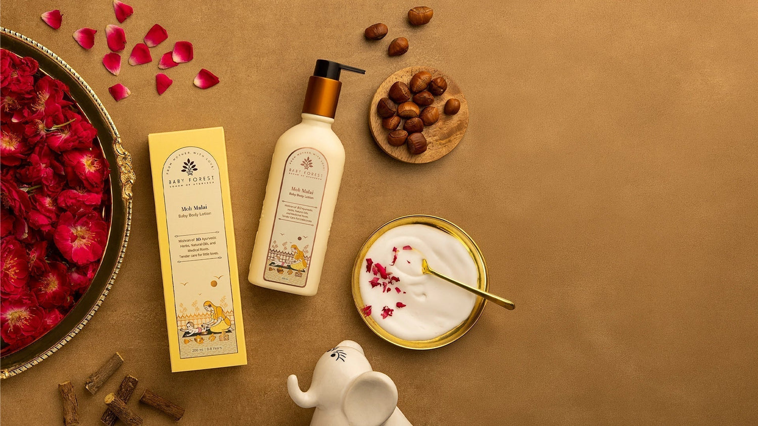 Baby Forest Moh Malai baby body lotion bottle and box with a bowl of creamy lotion, rose petals, and nuts on a brown background.