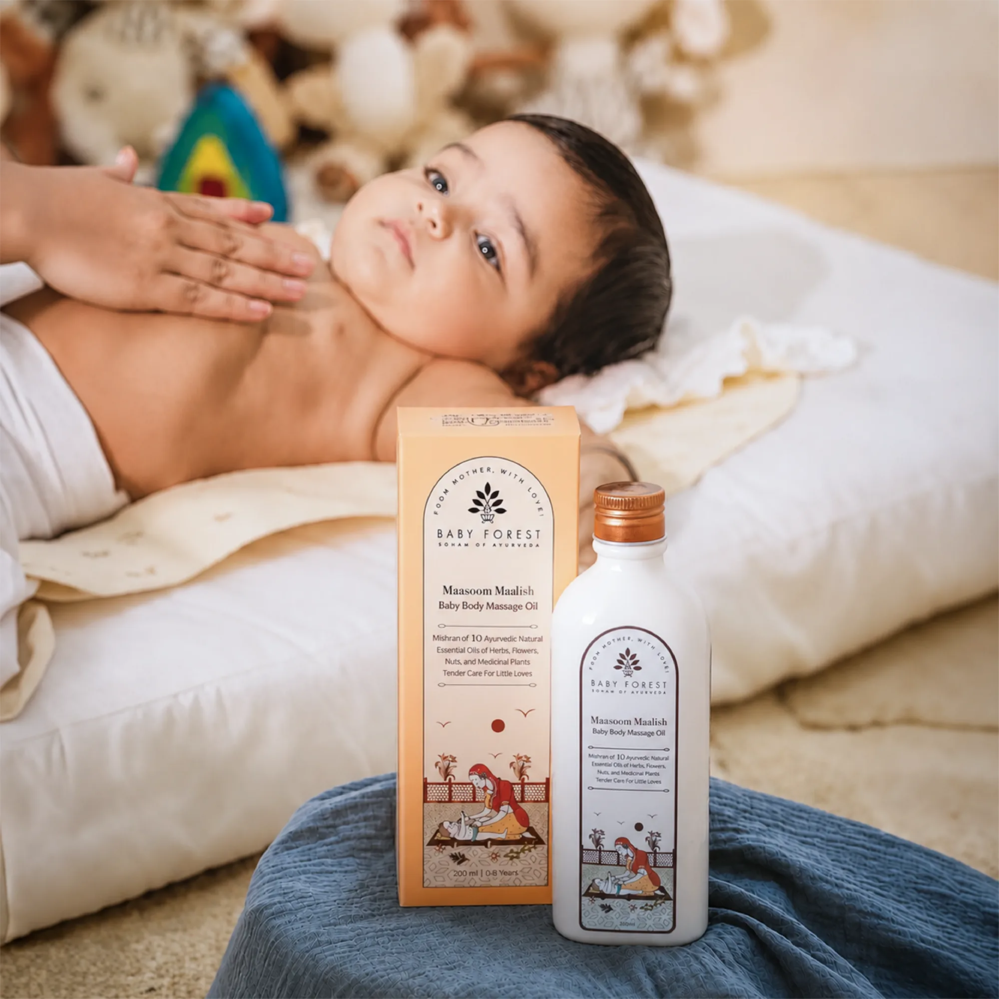 A baby lying on a white cushion while an adult massages the baby's chest. In the foreground, a bottle and box of Baby Forest Maasoom Maalish Baby Body Massage Oil are displayed on blue fabric.