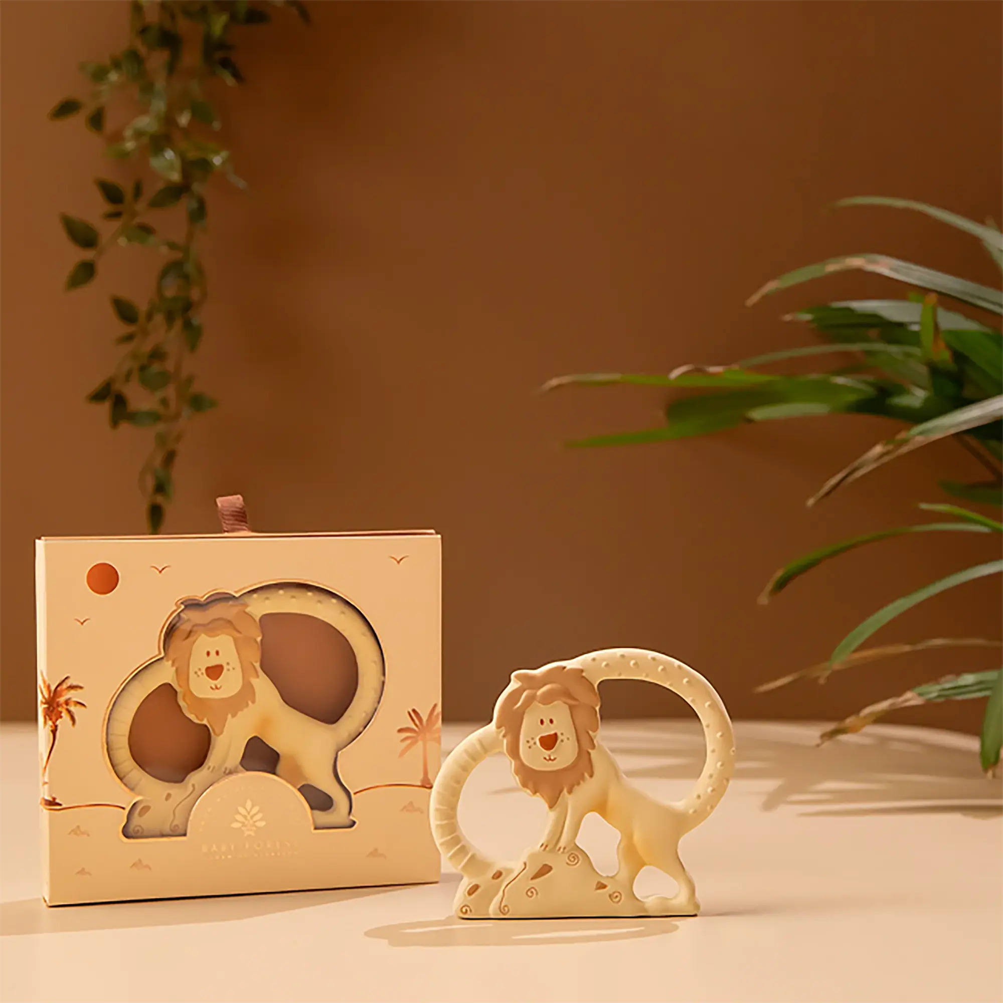 A cream-colored Natkhat Sher Natural Rubber Baby Teether displayed next to its matching decorative packaging box on a warm beige surface, with a warm brown background and soft green plant leaves.