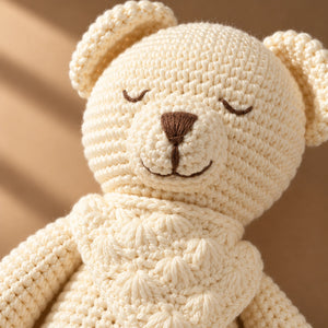 Close-up of a cream-colored crocheted teddy bear soft toy with embroidered closed eyes, a brown nose, and a stitched smile, set against a warm beige background.