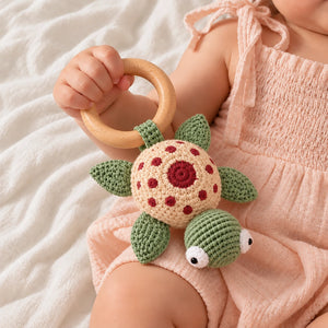 A baby in a pink smocked romper holds a crocheted turtle rattle with a wooden ring toy against a soft white textured blanket background. - Crochet Karigari Cuddly Teddy Toy