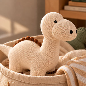A knitted Cuddly Buddies Baby Dino Soft Toy in a warm cream color with small black eyes and a brown textured back, resting inside a matching cream fabric basket with striped fabric nearby.