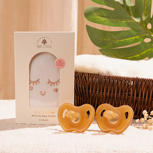 Two Sneh Dulaar Baby Pacifier in front of their packaging with a wicker basket and green leaves in the background.