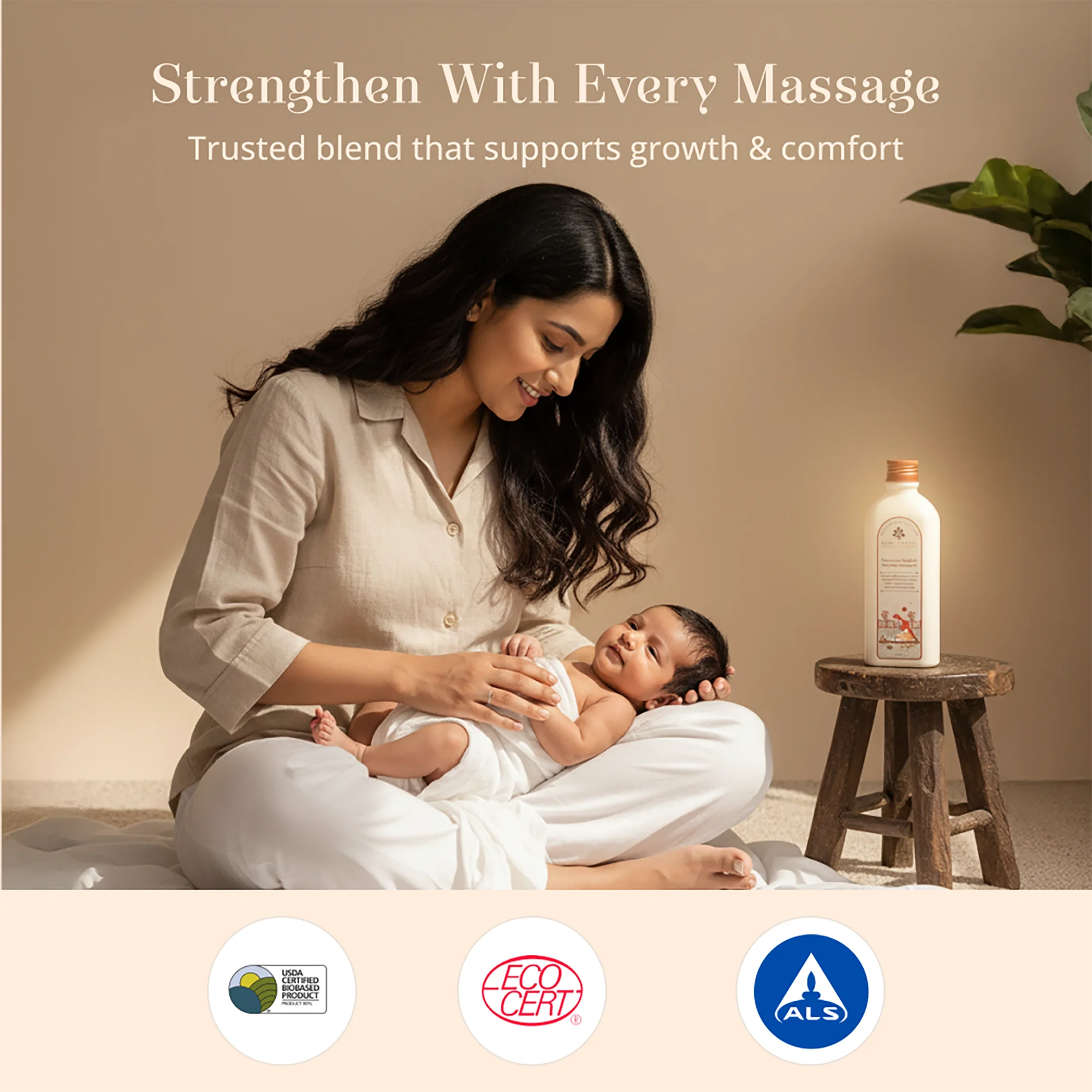 A woman in a beige shirt and white pants gently massages a baby lying on her lap in a warm, softly lit room. A small wooden stool beside her holds a Maasoom Maalish Baby Body Massage Oil.