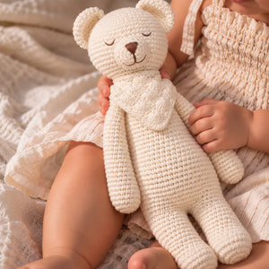 A close-up lifestyle shot of a baby in a cream-colored smocked dress holding a handmade cream crocheted teddy bear.- Crochet Karigari Cuddly Teddy Toy