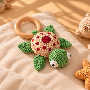 Crocheted turtle baby toy with a wooden ring handle, resting on a beige blanket with warm sunlight and shadows.