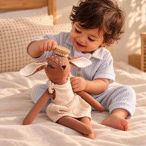 A toddler in light blue striped pajamas sits on a cream bed, gently brushing a brown plush Cuddly Buddies Baby Deer Soft Toy with a wooden soft brush.