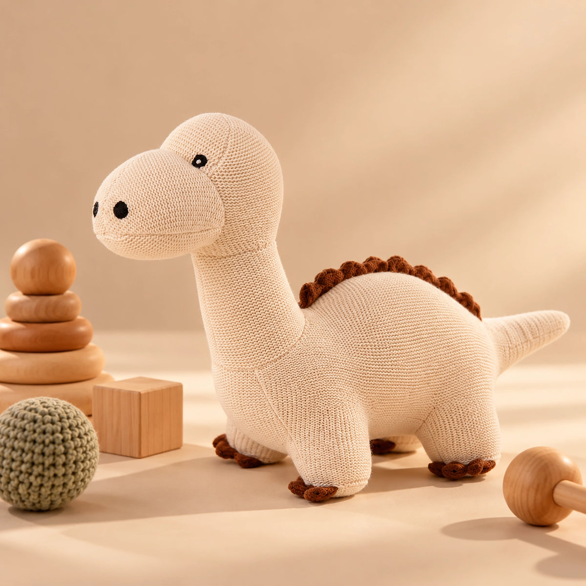 A knitted Cuddly Buddies Baby soft dinosaur toy in cream with brown accents, styled on a warm beige surface with wooden stacking toys and a small crocheted ball.