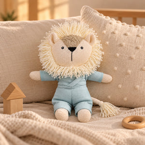 A Cuddly Buddies Baby Simba Soft Toy with a cream-colored yarn mane, wearing a light blue outfit, sitting on a cozy beige blanket with a wooden house toy and a wooden ring teether nearby.