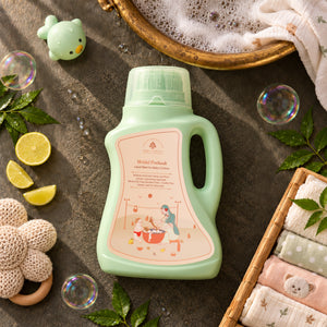 Mridul Poshaak - Liquid Wash for Baby’s Clothes with a pink label, styled in a flat-lay arrangement with baby towels, soap bubbles, lemon slices, and a gold dish on a dark textured surface.