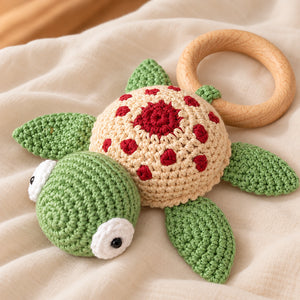 Crocheted turtle baby toy with a wooden ring teether, featuring a green head and flippers, a beige shell with red polka dots and a red center, and white eyes, resting on a beige fabric background.
