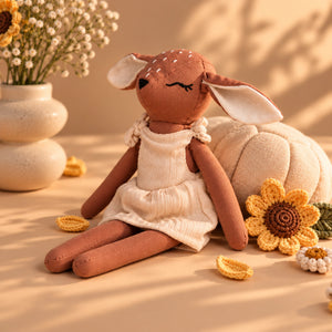 A Cuddly Buddies Baby Deer Soft Toy with long floppy ears, a brown body, and a cream dress sits on a warm beige surface with decorative sunflowers, a pumpkin, and small flowers in ceramic vases.