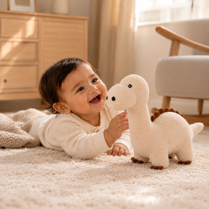 A baby lying on a cream carpet in a bright room, smiling at a Cuddly Buddies Baby Dino Soft Toy. The baby wears a cream long-sleeve top, and a light wood dresser and chair are visible in the background.