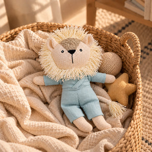 A Cuddly Buddies Baby Simba Soft Toy with a cream-colored yarn mane, wearing light blue overalls, resting in a woven wicker basket lined with beige textured blankets. A small yellow star toy is tucked beside it.