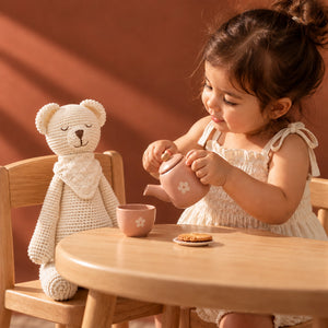 A toddler girl in a cream dress sits at a wooden table, pouring tea from a pink teapot into a pink cup. A knitted Crochet Karigari Cuddly Teddy Toy sits on the chair beside the table.