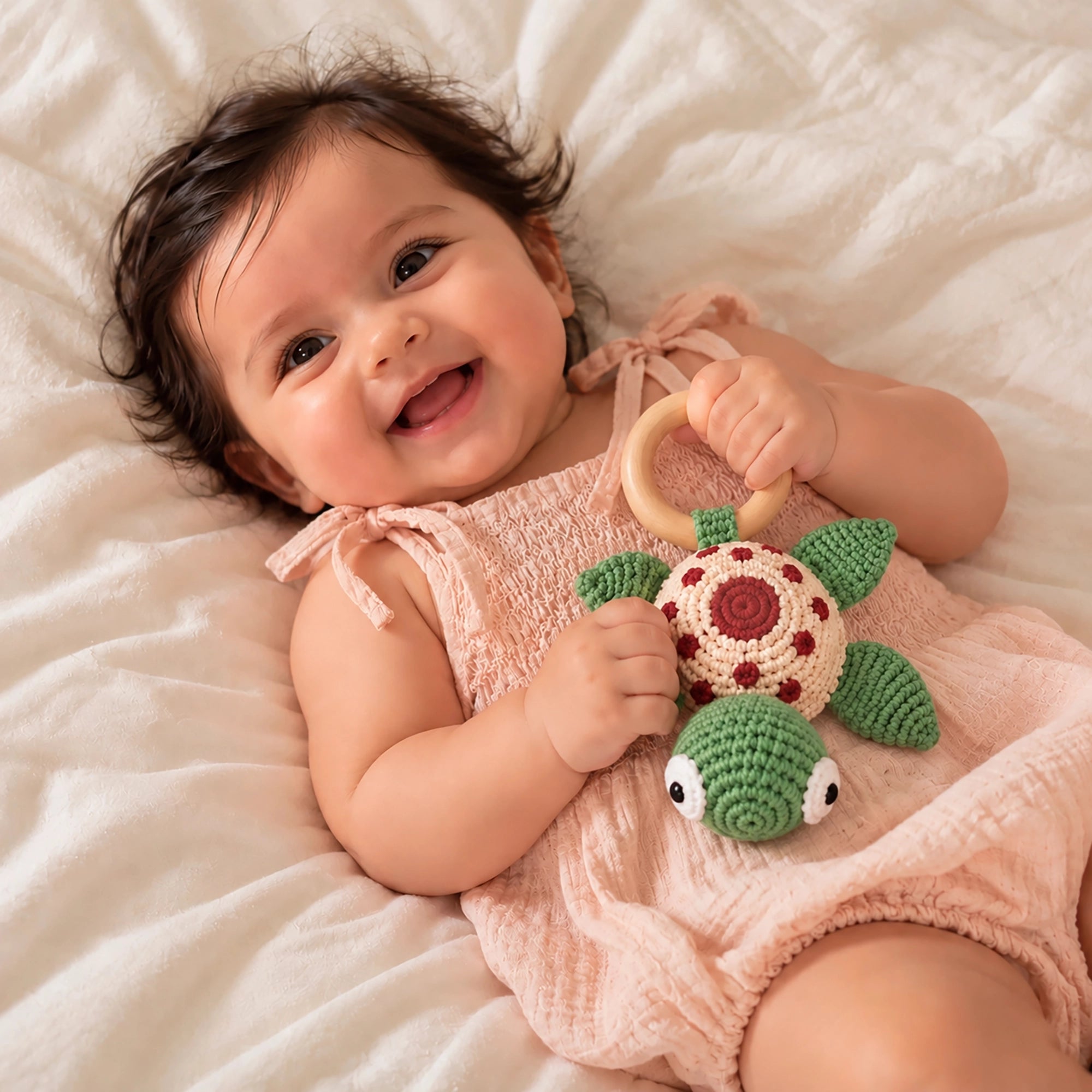 A smiling baby lying on a white textured blanket, wearing a pale pink sleeveless romper and holding a Crochet Karigari Cuddly Teddy Toy with a wooden ring.