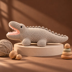 A Cuddly Buddies Baby Croco Soft Toy with an open mouth, displayed on a round pedestal with a ball of yarn, wooden beads, and a stack of wooden rings against a warm brown background.