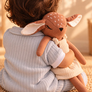 A child wearing a light blue ribbed long-sleeve top holds a Cuddly Buddies Baby Deer Soft Toy with a brown body, cream inner ears and dress, and small white spots on its forehead, in a warm indoor setting.