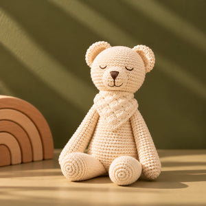 A cream-colored crocheted teddy bear soft toy with a matching knitted scarf, sitting on a wooden surface against an olive green background with soft diagonal shadows.