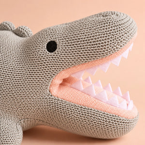 Close-up of a Cuddly Buddies Baby Croco Soft Toy in warm gray, with a peach-pink interior, white teeth, and a black eye on a soft peach background.