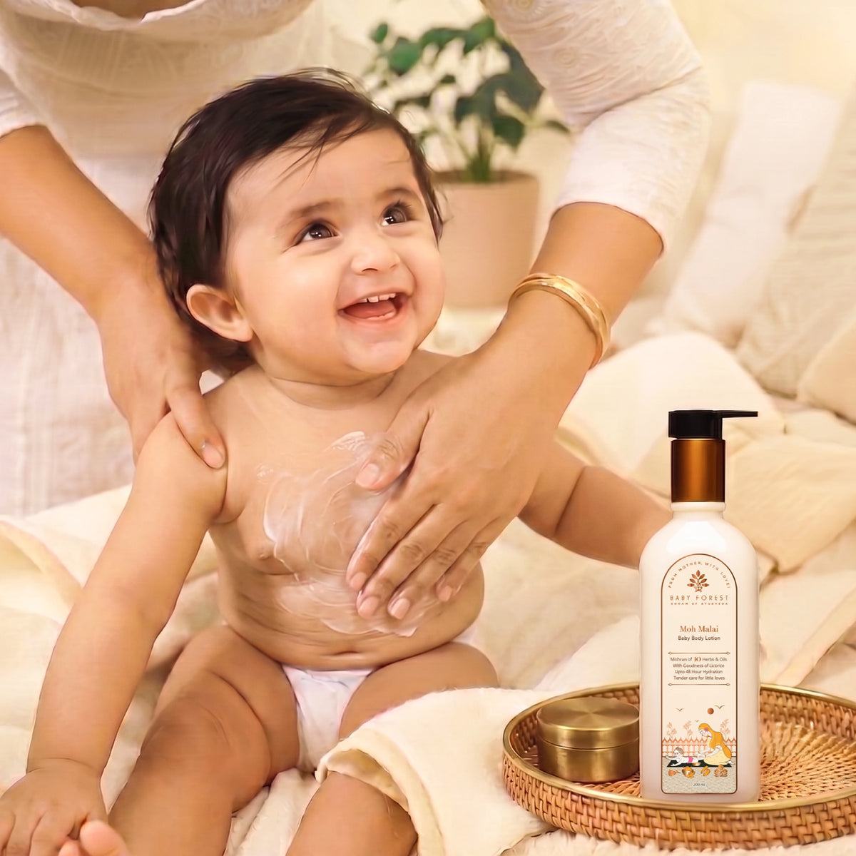 A baby is sitting on a cream-colored bed while an adult applies Moh Malai Baby Body Lotion to the baby's chest.