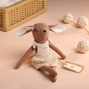 A beige-colored baby deer soft toy with white spots on its head, dressed in a tiny outfit, sitting next to decorative items on a pink background.