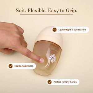 A hand holds a soft, flexible baby bottle with a translucent beige body and a cream-colored cap. The image features text callouts highlighting its key features: 'Soft. Flexible. Easy to Grip.'