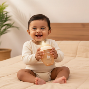 A smiling baby sitting on a beige quilted bed holding a Laad Pyaar Baby Feeding Bottle with a white cap. 