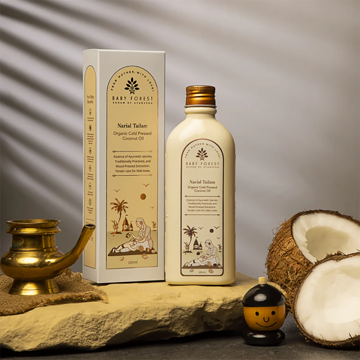A Narial Tailam- Organic Cold Pressed Coconut Oil are displayed on a stone slab with decorative props, including a brass lamp, a small smiley face jar, and halved coconuts, against a gray background with leaf shadows.