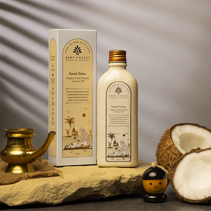A Narial Tailam- Organic Cold Pressed Coconut Oil are displayed on a stone slab with decorative props, including a brass lamp, a small smiley face jar, and halved coconuts, against a gray background with leaf shadows.