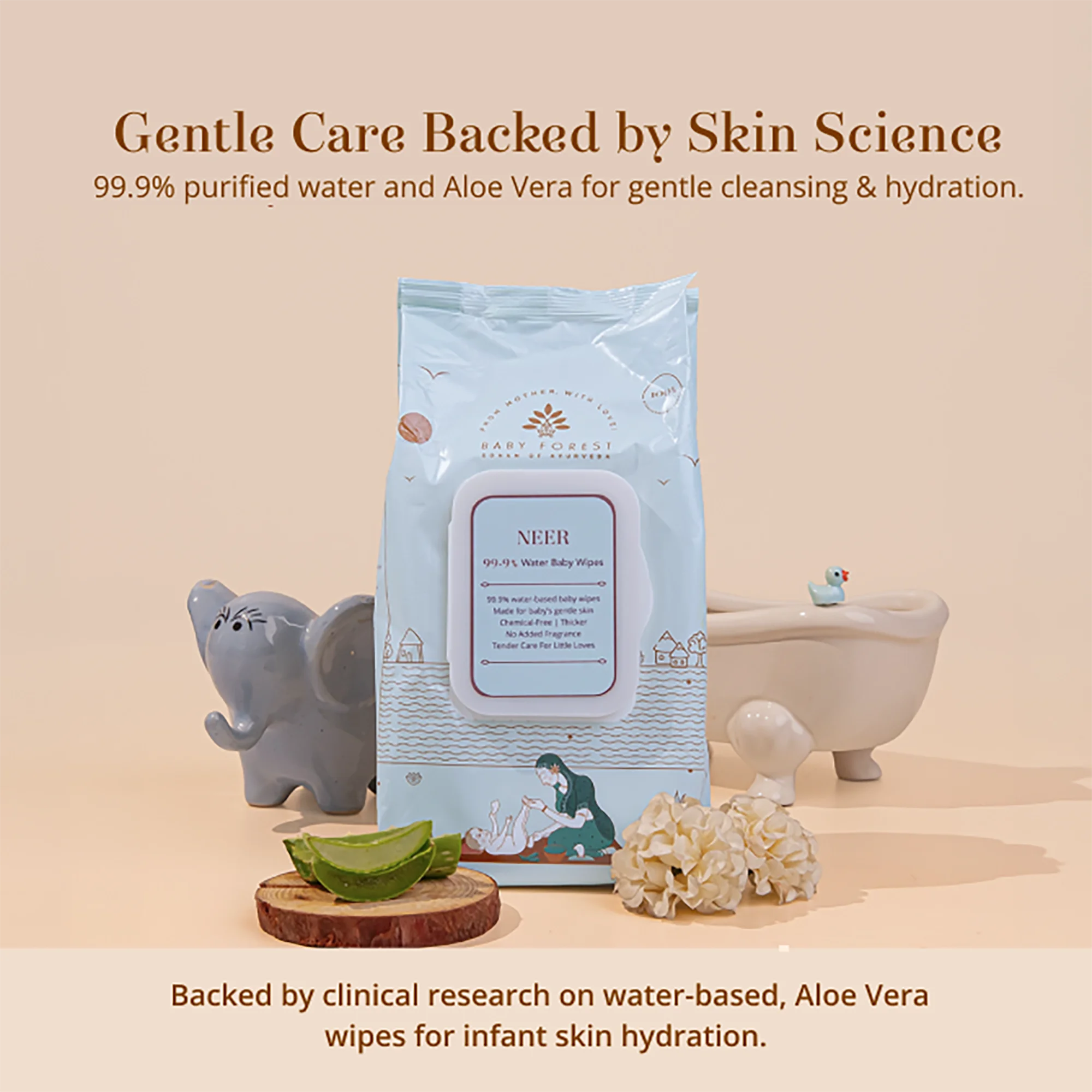 A package of Neer 99.9% Water Baby Wipes stands upright on a beige studio background, surrounded by baby-care props including a gray elephant figurine, aloe vera slices, a white claw brush, and a small bath tub prop.
